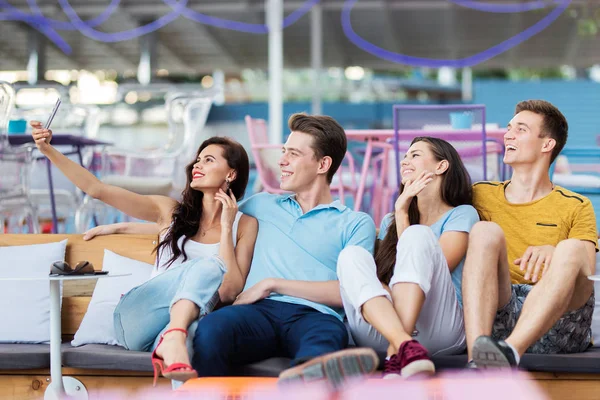 Eine Gruppe gut aussehender Freunde sitzt lachend mit Kissen auf der Bank und macht Selfies im netten Sommercafé. Unterhaltung, Spaß haben. Freundschaft. — Stockfoto