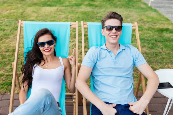 Um belo casal nos óculos de sol está deitado nas cadeiras do convés no gramado no agradável café de verão. Entretenimento, divertindo-se. Amizade, relação . — Fotografia de Stock