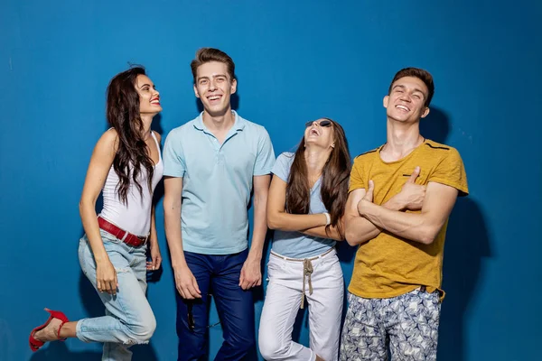 Four good-looking friends are laughing while standing in front of the blue wall having confident and happy looks. Entertainment, having good time. Friendship, relationship.