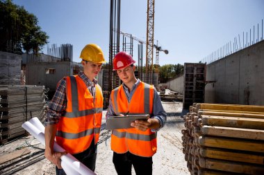 Mimar ve inşaat mühendisi turuncu iş yeleği giymiş ve kask bir bina proje sitesi İnşaat malzeme ile açık hava tablete tartışmak