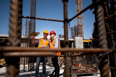 Mimar ve inşaat mühendisi turuncu yelek çalışmak ve kask bir bina proje sitesi çelik kare bir sürü bina açık hava üzerinde tartışmak