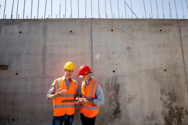 İnşaat mühendisi ve mimar turuncu iş yeleği giymiş ve kask bir beton duvar arka plan üzerinde durmak ve tablet kullanmak