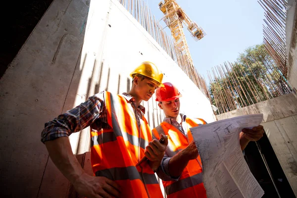 Deux ingénieurs civils vêtus de gilets de travail orange et de casques explorent la documentation du projet sur le chantier — Photo