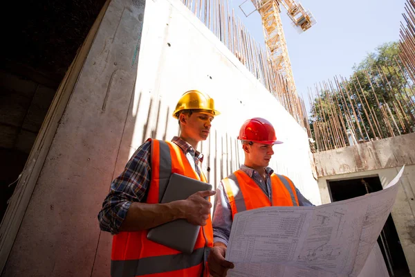 Deux ingénieurs civils vêtus de gilets de travail orange et de casques explorent la documentation du projet sur le chantier — Photo