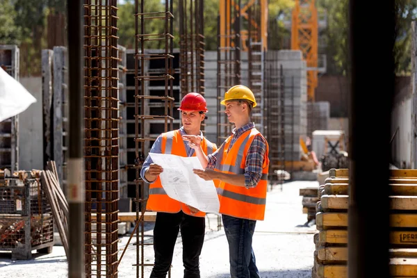 Direttore edile e ingegnere vestito con giubbotti di lavoro arancioni e caschi rigidi esplorare la documentazione di costruzione sul cantiere vicino ai telai in acciaio — Foto Stock