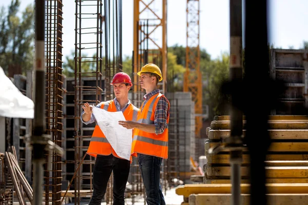 Le directeur de la construction et l'ingénieur vêtus de gilets de travail orange et de casques durs explorent la documentation de construction sur le chantier près des cadres en acier — Photo