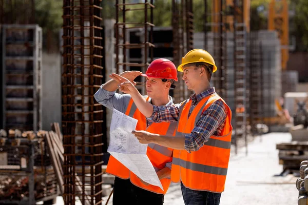 Ingénieur structurel et responsable de la construction en gilets de travail orange et casques durs explorent la documentation de construction sur le chantier près des cadres en acier — Photo