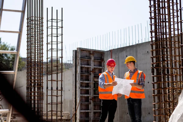 Civil engineer and construction manager in orange work vests and hard helmets explore construction documentation on the building site near the steel frames