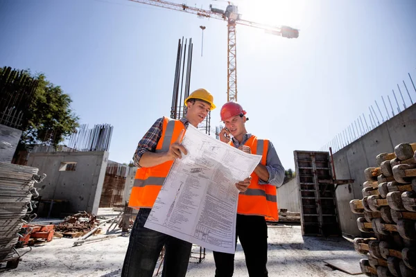 Ingegnere strutturale e responsabile della costruzione in giubbotti di lavoro arancioni e caschi rigidi discutere la documentazione e parlare per telefono sul cantiere all'aperto — Foto Stock