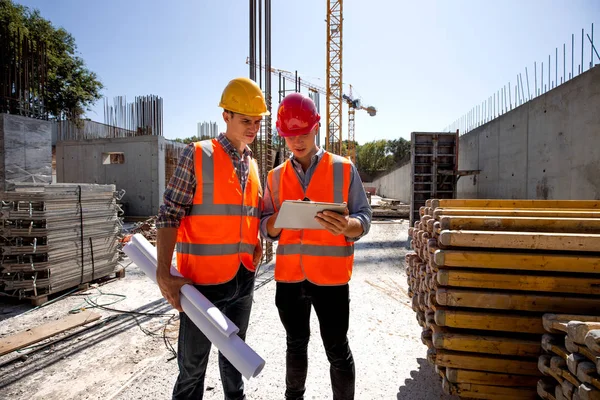 Architetto civile e responsabile della costruzione vestito con giubbotti e caschi arancioni discutono un progetto di costruzione sul tablet mobile sul cantiere aperto accanto alla gru — Foto Stock