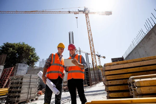 Civila arkitekt och bygg manager klädd i orange arbete västar och hjälmar diskutera ett byggprojekt på mobila tabletten på webbplatsen öppen byggnad bredvid kranen — Stockfoto