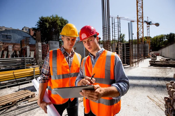 Architect  and structural engineer dressed in orange work vests and  helmets discuss a building project on the tablet on the open air building site with construction material
