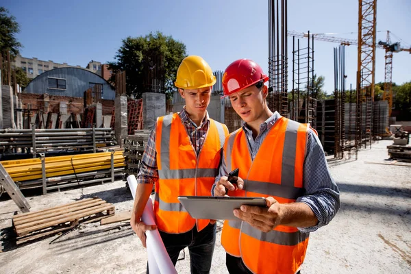 Architect  and structural engineer dressed in orange work vests and  helmets discuss a building project on the tablet on the open air building site with construction material — Stock Photo, Image