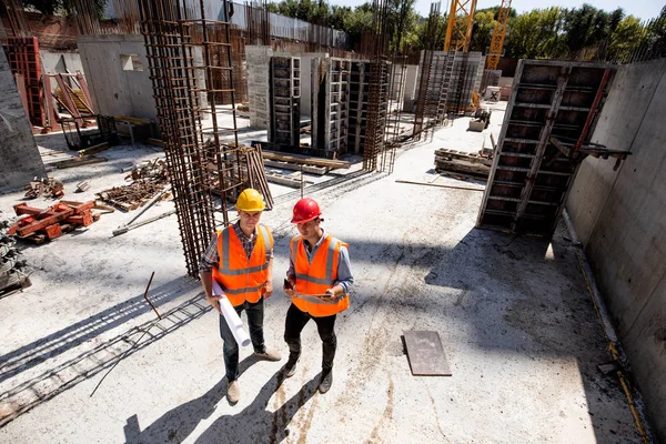 Ingegnere civile e architetto vestito con giubbotti arancioni e pipistrelli duri discutere il processo di costruzione sul cantiere aperto con materiale da costruzione — Foto Stock