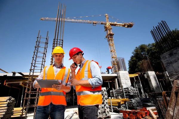 Ingegnere strutturale e architetto vestito con giubbotti di lavoro arancioni e cappelli duri discutere il processo di costruzione al telefono e utilizzare tablet sul cantiere aperto — Foto Stock