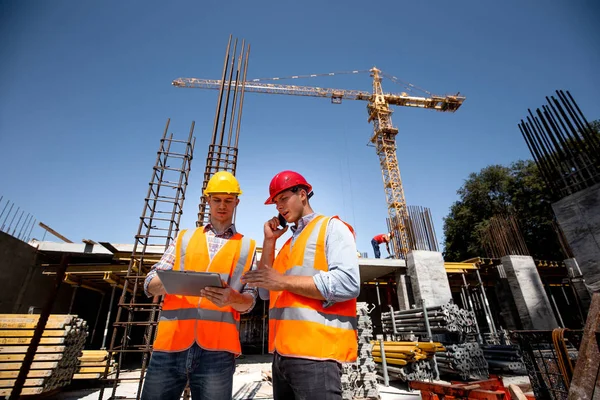 Structural engineer and architect dressed in orange work vests and  hard bats  discuss the construction process by the phone and use tablet  on the open building site near the crane — Stock Photo, Image