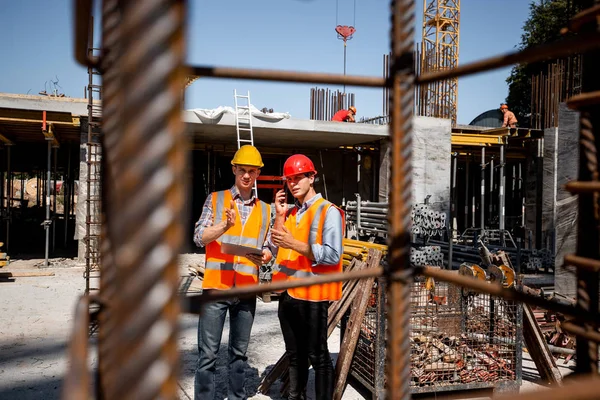 Architect  and structural engineer  in orange work vests and  helmets discuss a building project on the open air building site with a lot of steel frames — Stock Photo, Image