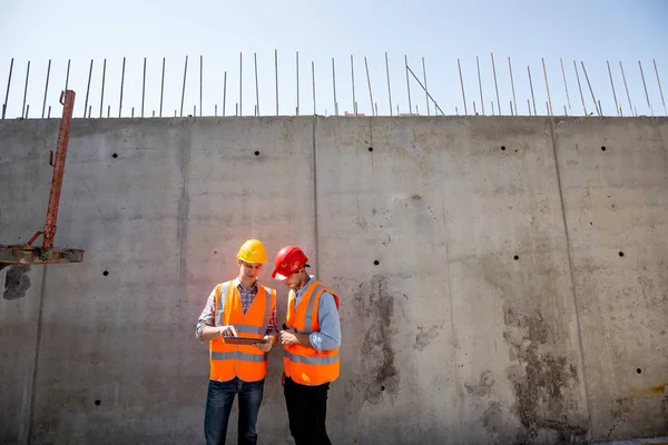 Civil engineer and architect dressed in orange work vests and  helmets stand on a concrete wall background and use tablet — Stock Photo, Image