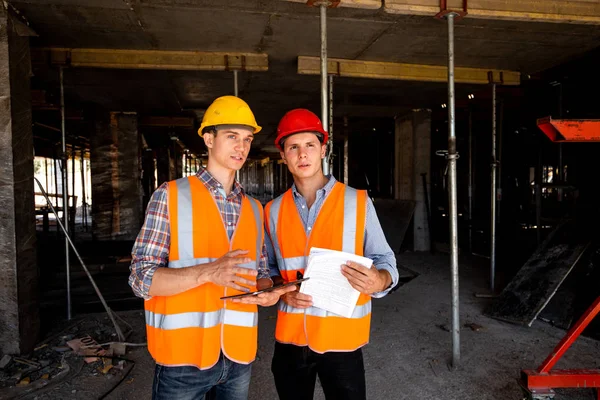 Two young civil engineers dressed in orange work vests and helmets work with tablet and  documentation inside the building under construction