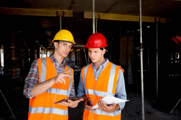 Constructeur en architect gekleed in oranje werk vesten en helmen tablet gebruiken en houden van de projectdocumentatie binnen het gebouw in aanbouw — Stockfoto