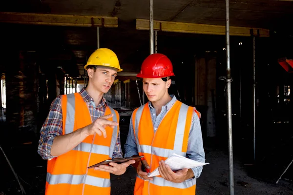 Ingénieur structurel et architecte vêtus de gilets de travail orange et casques utilisent une tablette et gardent la documentation du projet à l'intérieur du bâtiment en construction — Photo