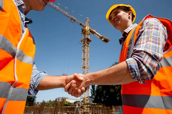 Två män klädda i orange arbete västar och hjälmar skaka hand på byggnadsplatsen nära kranen — Stockfoto
