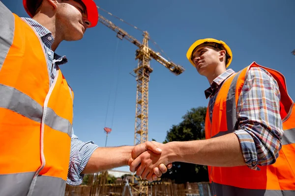 Två män klädda i orange arbete västar och hjälmar skaka hand på byggnadsplatsen nära kranen — Stockfoto