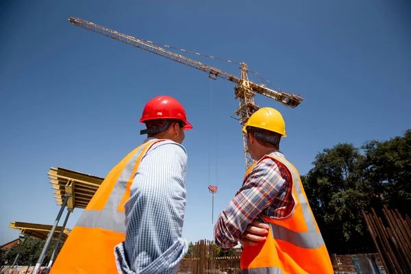 Two men dressed in orange work vests and  helmets look at th crane on the building site — Stock Photo, Image