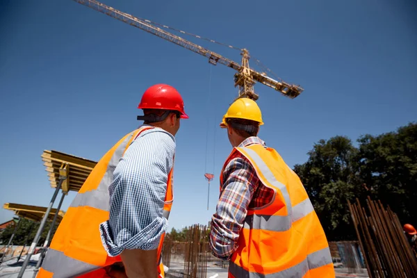 Two men dressed in orange work vests and  helmets look at th crane on the building site — Stock Photo, Image