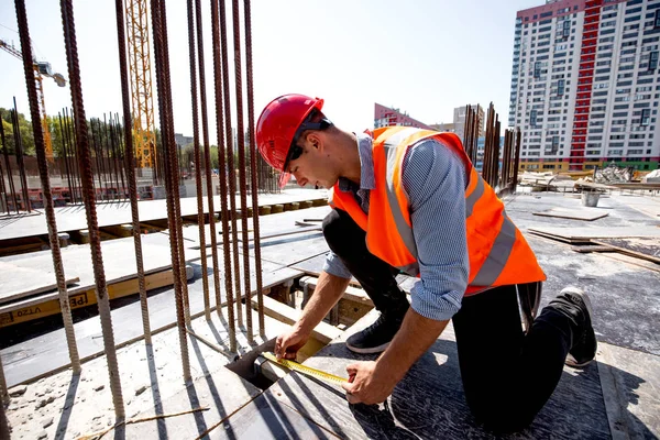 Civil engineer dressed in shirt, orange work vest and helmet measures the hole with a tape measure on the building site — Stock Photo, Image