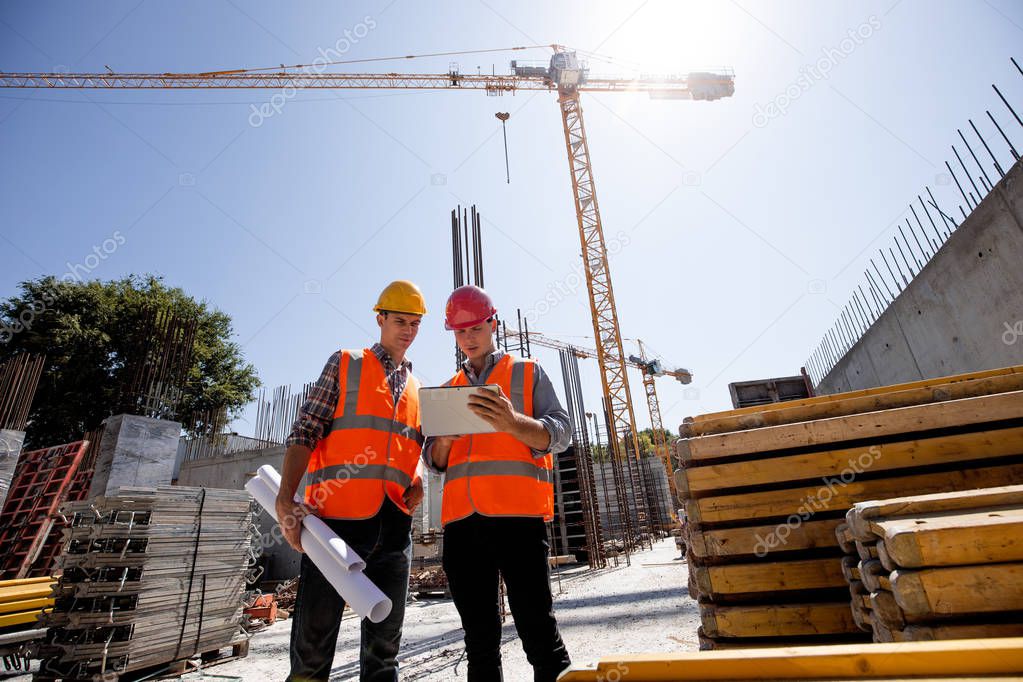 Civil architect  and construction manager  dressed  in orange work vests and  helmets discuss  a building project on the mobile tablet on the open building site next to the crane