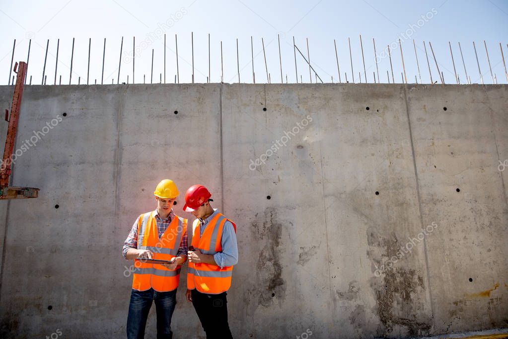 Civil engineer and architect dressed in orange work vests and  helmets stand on a concrete wall background and use tablet