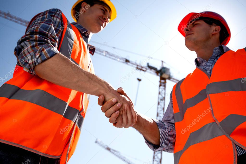 Structural engineer and architect dressed in orange work vests and  helmets shake hands on the building site near the crane