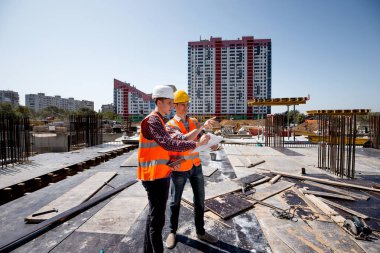 Mimar ve inşaat Yöneticisi turuncu iş yeleği giymiş ve kask belgeleri sitenin vinç yanındaki bina açık hava üzerinde tartışmak