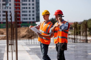İki yapısal mühendisleri gömlekler, turuncu iş yeleği giymiş ve kask inşaat dokümantasyon keşfetmek ve şantiye çelik çerçeve yakınındaki telefonla konuşmak