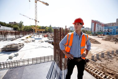 İnşaat alanında bir çok katlı bina arka planı üzerinde gömlek, turuncu iş yelek ve kask giymiş genç adam duruyor