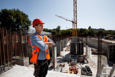 İnşaat alanında bir çok katlı bina arka planı üzerinde gömlek, turuncu iş yelek ve kask giymiş genç adam duruyor