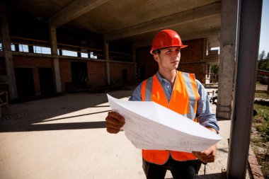 Giyinmiş genç adam gömlek, turuncu iş yelek ve kask binanın yapım aşamasında içinde bina sitede inşaat dokümantasyon keşfetmek