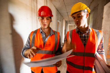 İki adam turuncu iş yeleği giymiş ve kask yapım aşamasında binada inşaat belgeleriyle çalışma