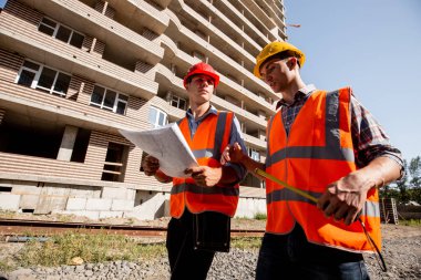 İki inşaat mühendisi gömlekler, turuncu iş yelek giymiş ve kask çok katlı bina yakınındaki yürümek