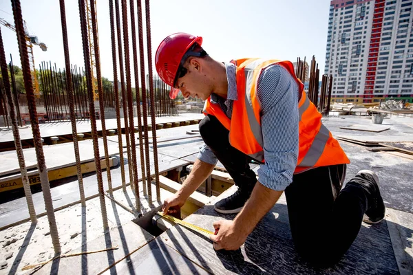 Man dressed in shirt, orange work vest and helmet measures the hole with a tape measure on the building site — Stock Photo, Image