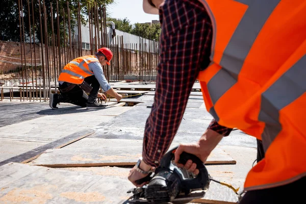 Builder dressed in orange work vest and helmet is using a circular saw on the open building site — Stock Photo, Image