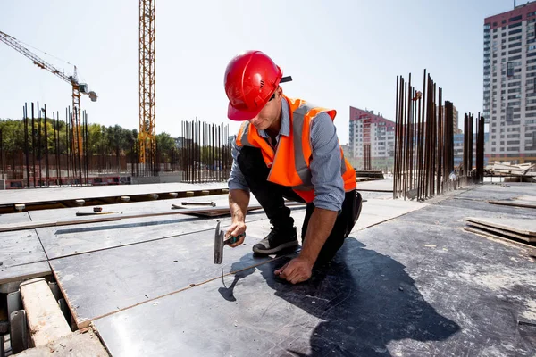 Builder dressed in orange work vest and helmet hammers a nail on the open building site — Stock Photo, Image