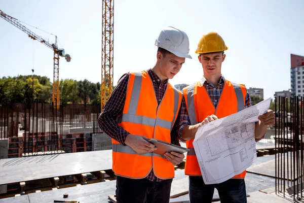 Structural engineer and construction manager dressed in orange work vests and helmets discuss documentation on the open air building site next to the crane — Stock Photo, Image