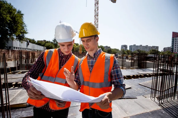 Structural engineer and construction manager dressed in orange work vests and helmets discuss documentation on the open air building site next to the crane — Stock Photo, Image