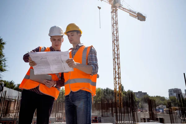 Architect en constructie manager gekleed in oranje werk vesten en helmen bespreken documentatie op de open lucht bouwplaats naast de kraan — Stockfoto