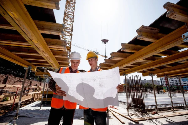 Deux hommes vêtus de chemises, gilets de travail orange et casques explorent la documentation de construction sur le chantier près des constructions en bois — Photo