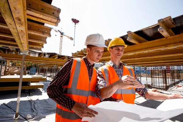 Two men dressed in shirts, orange work vests and helmets explore construction documentation on the building site near the wooden building constructions — Stock Photo, Image