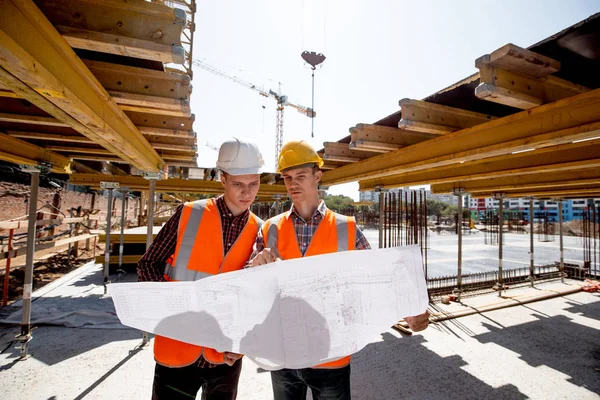 Dois homens vestidos com camisas, coletes laranja e capacetes exploram a documentação de construção no local de construção perto das construções de edifícios de madeira — Fotografia de Stock