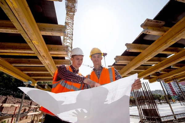 Structural engineer and architect dressed in shirts, orange work vests and helmets explore construction documentation on the building site near the wooden building constructions — Stock Photo, Image